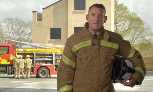 Fire officer wearing dark mustard jacket and yellow high vis standing in front of a red fire engine