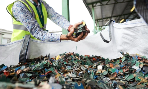 a pale skinned man in a hi-viz jacket standing next to a chute with shredded electricals with his hand outstretched to catch some