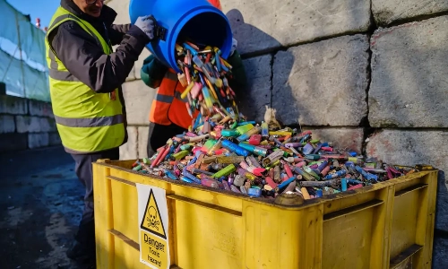 a mid shot of two men emptying a blue barrel of used single use vapes into a large yellow container