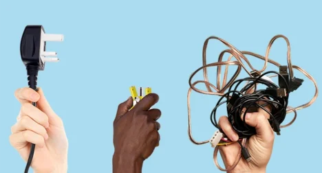 Two light-skinned hands and one dark skinned one reaching up from the bottom of the image, one holding a yellow electrical cable the other holding black USB cables and the third holding a cable with a three pin plug on against a pale blue backdrop.
