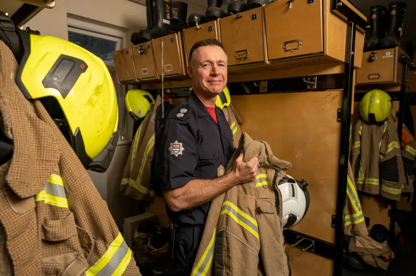 Station officer Shawn Howlett from London Fire Brigade in the kit room of Euston fire station holding his uniform and smiling to the camera