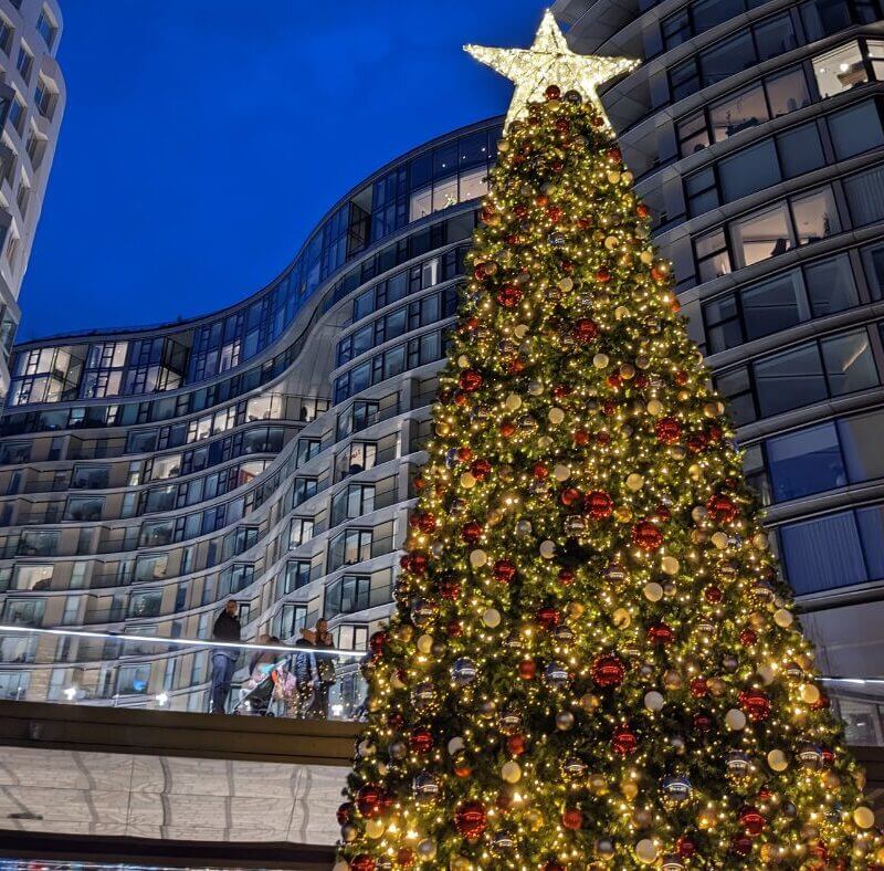 A very tall christmas tree decorated in bright yellow and white lights, topped with a light up star with buildings in the background